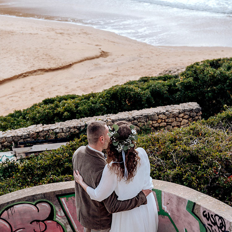Brautpaar mit Blumenkranz blickt auf den Strand der Algarve nach der freien Trauung